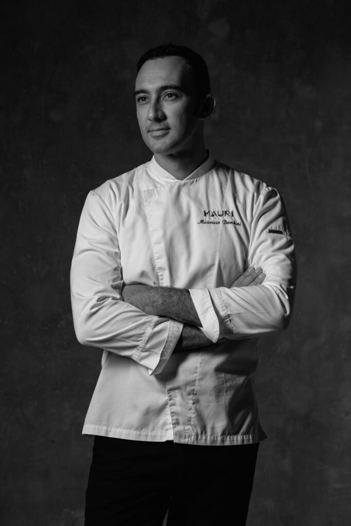 Black and white portrait of a chef in white uniform with arms crossed, name embroidered on chest.